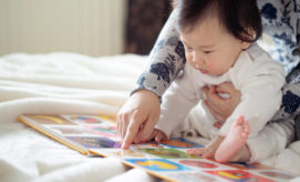 A baby, supported by an adult's arm, attentively looks at an open picture book on a soft, white bed, while the adult points at colorful images on the pages.