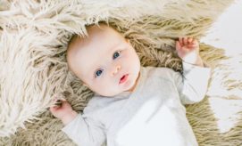 A baby lies on a soft, fluffy beige rug, gazing upward with wide eyes and wearing a light gray onesie, surrounded by gentle sunlight.