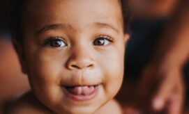 A smiling baby with bright eyes and tongue out sits indoors, surrounded by soft lighting and blurred background, conveying a joyful and playful atmosphere.