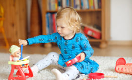 A young child, wearing a blue dress with owl prints, plays with a doll seated in a red high chair on a carpet, surrounded by toy dishes in a cozy room.