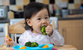A toddler sits in a high chair, eating broccoli from a blue plate with orange slices, in a kitchen with wood cabinets and a tiled backsplash.