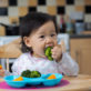 A toddler sits in a high chair, eating broccoli from a blue plate with orange slices, in a kitchen with wood cabinets and a tiled backsplash.
