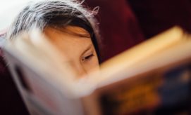 A child reads a book intently, partially obscured by its pages, in a cozy setting with a blurred, dark red background. No text is visible on the book cover.