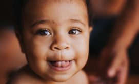 A baby smiles with their tongue out, looking directly at the camera. In the background, a blurred figure offers a warm, intimate setting.