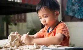 A young child intensely sculpts a clay model with a small tool while seated at a wooden table, surrounded by a cozy interior with blurred furniture and plants in the background.