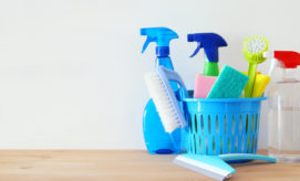 Cleaning supplies, including spray bottles, sponges, a brush, and a scrubber, are organized inside and around a blue basket. They rest on a wooden surface against a plain white background.