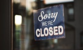 A blue sign with white and red text reads "Sorry we're CLOSED," hanging on a glass door, indicating the establishment is not open. The background shows a blurred interior.
