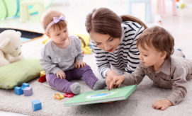 An adult and two toddlers lie on a carpeted floor, reading a book with a green cover in a playroom, surrounded by colorful toys and a teddy bear.