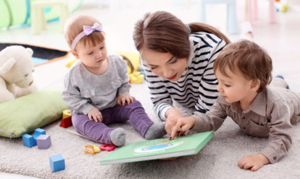 An adult and two toddlers lie on a carpeted floor, reading a book with a green cover in a playroom, surrounded by colorful toys and a teddy bear.
