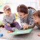 An adult and two toddlers lie on a carpeted floor, reading a book with a green cover in a playroom, surrounded by colorful toys and a teddy bear.