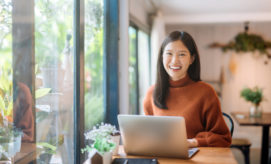 A person smiles while sitting at a table with a laptop, surrounded by potted plants, near large windows, in a cozy, well-lit space.