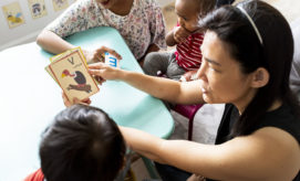Adults and children sit around a table engaging in a learning activity. One adult holds a flashcard displaying a vulture, marked with the letter "V," while the others observe attentively.