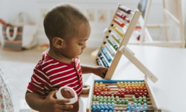 A child in a red striped shirt plays with a colorful abacus, holding a small ball, in a bright, cozy room with a striped bag in the background.