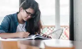 A person with long hair and glasses writes in a notebook while sitting at a table, surrounded by a bright, softly lit room with large windows.