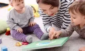 A woman reads a green book with two toddlers, engaging them in a cozy living room setting with colorful toys scattered around.