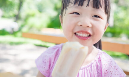 A child smiles while holding a popsicle, sitting on a bench in a sunny park with blurred greenery in the background.