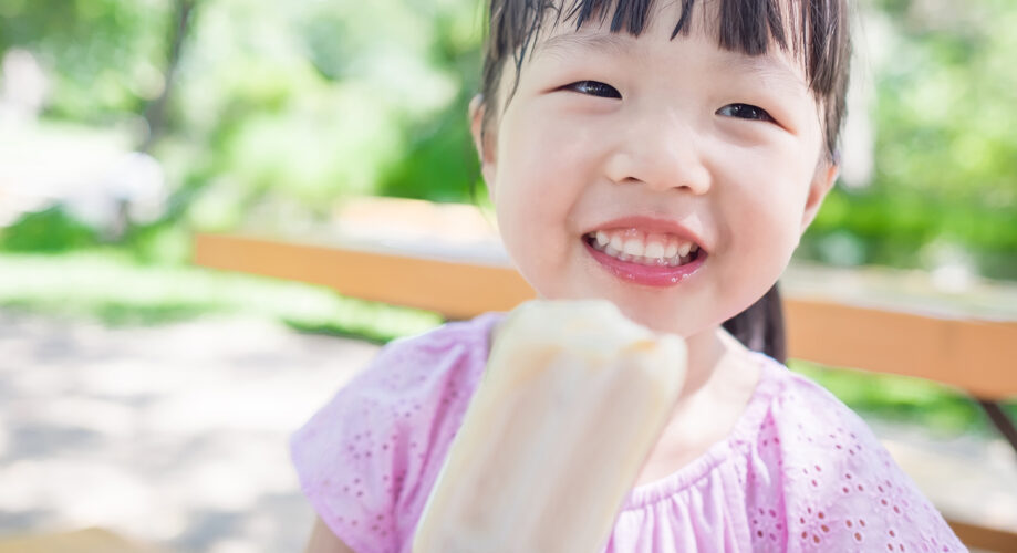 A child smiles while holding a popsicle, sitting on a bench in a sunny park with blurred greenery in the background.