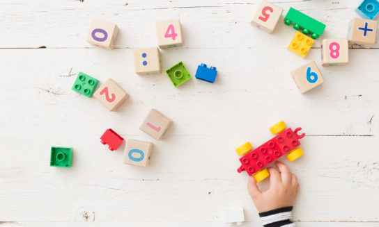 A child's hand is assembling a red and yellow toy car with building blocks, surrounded by numbered wooden cubes and additional colorful blocks on a white wooden surface. Visible numbers: 0, 4, 2, 6, 8, 9.