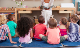 A teacher reads a book to children sitting on blue mats in a classroom, surrounded by educational materials and plants on shelves.