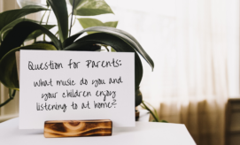 Card displayed on a plant holder, reading: "Question for Parents: What music do you and your children enjoy listening to at home?" Set against a backdrop of indoor plants and soft window light.