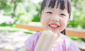 A child smiles while holding a popsicle, sitting on a bench in a sunny park with blurred greenery in the background.