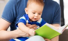 A baby sits on an adult's lap, holding and looking at a green book, in a cozy home setting. The adult supports the baby, dressed in a blue shirt.