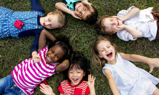 Children lie on grass, forming a circle with their heads together, smiling and laughing, wearing colorful clothing. The outdoor setting suggests a playful, joyful atmosphere.