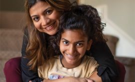 A woman embraces a smiling child from behind, both looking at the camera. They are sitting indoors, near a staircase, creating a warm and cozy setting.