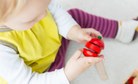 A toddler holds a red, segmented wooden apple toy, pulling it apart. The child wears a yellow shirt and purple striped pants, sitting on a light-colored floor.