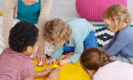 Children are drawing with crayons on a large yellow sheet, supervised by an adult. They are sitting on a patterned rug, surrounded by colorful cushions in a bright room.