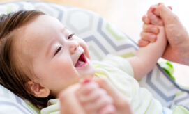 A baby laughs joyfully, holding an adult's hands. The infant is seated on a patterned cushion, creating a cozy and playful environment.