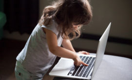 A young child, standing and focused, presses keys on a laptop placed on a table in a dimly lit room, concentrating intently on the keyboard.