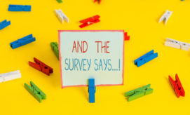 A paper sign held by a blue clothespin reads "AND THE SURVEY SAYS...!" surrounded by scattered colorful clothespins on a bright yellow background.