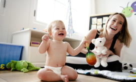 A baby joyfully shakes a colorful maraca while sitting on the floor. Nearby, a smiling adult holds a white teddy bear in a bright, toy-filled playroom.