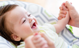 Baby smiles widely while lying in a geometric-patterned chair, holding an adult's hands. Bright, soft lighting creates a cheerful and affectionate atmosphere.