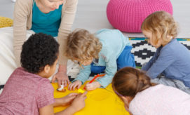 Children draw on a yellow paper on the floor, supervised by an adult. They are surrounded by colorful cushions and a patterned rug, creating a playful, engaging environment.