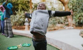 A child is joyfully jumping with arms outstretched in a garden, surrounded by green grass and trees, while people stand in the background.