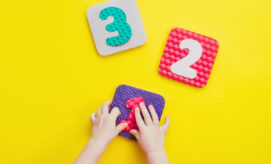 Child's hands manipulate a square tile featuring the number "1" on a bright yellow surface. Nearby, additional tiles display the numbers "2" and "3."