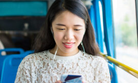 A woman wearing earphones is smiling while looking at her phone. She sits on a blue seat inside a bus, with a window showing a blurred view of the outside.