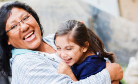 A woman and a young girl are joyfully embracing, both smiling broadly in an outdoor setting. The woman wears glasses and a plaid shirt, while the girl has her hair in a ponytail.
