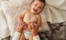 A smiling baby lies on a soft, textured blanket. Adult hands gently hold and tickle the baby's sides. The background includes plush, gray and white fabrics.