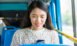 A woman listens to music on earphones while looking at her phone, seated on a blue bus. The background shows bus interiors and a blurred outside view.