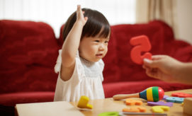 A child wearing a white dress raises a hand while looking at a red number "5" held by another person, surrounded by colorful toys on a wooden table in a cozy room.