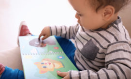 A toddler, wearing a striped shirt, is seated and exploring a colorful animal book featuring illustrations of animals in a playful, home environment.
