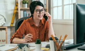 A woman, speaking on the phone, sits at a desk with architectural plans, pencils, and coffee cups in a bright office with shelves and a clock in the background.