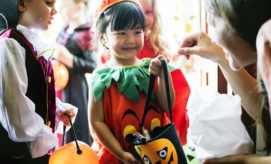 Child in a pumpkin costume receives candy from an adult during a Halloween trick-or-treating event, surrounded by other children in costumes holding treat bags.