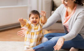 A baby, wearing a yellow plaid dress, smiles and reaches out while sitting on a carpeted floor. A woman beside her, in a gray cardigan, looks at her warmly within a cozy living room.