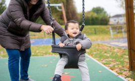 A child, smiling in a swing, is gently pushed by an adult wearing a brown coat. They are in a playground with fallen leaves on the grass and trees in the background.