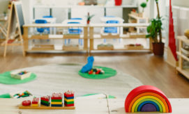 Colorful wooden toys sit on a table in a bright classroom, with a rainbow stacker and various blocks, surrounded by child-sized furniture, blue chairs, and plants.