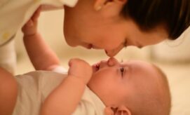A baby lies on its back, looking up, while a woman leans over, gently touching noses in a warm, intimate setting.